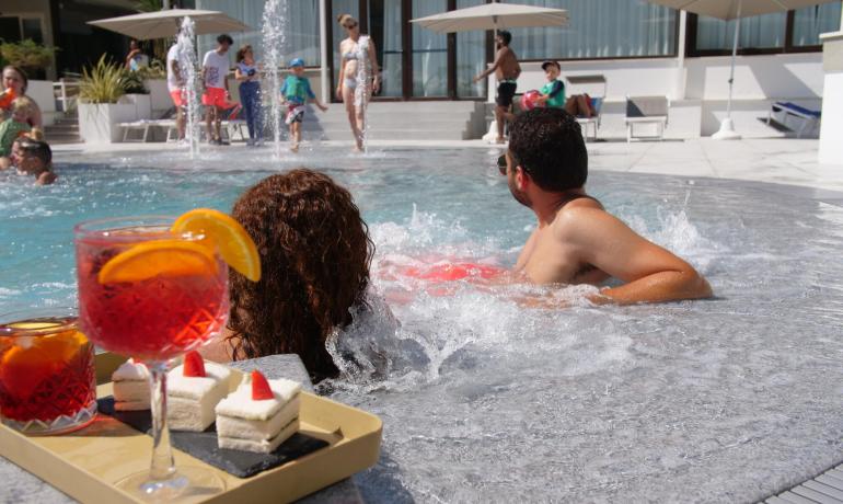 Persone si rilassano in piscina con drink e dolci.
