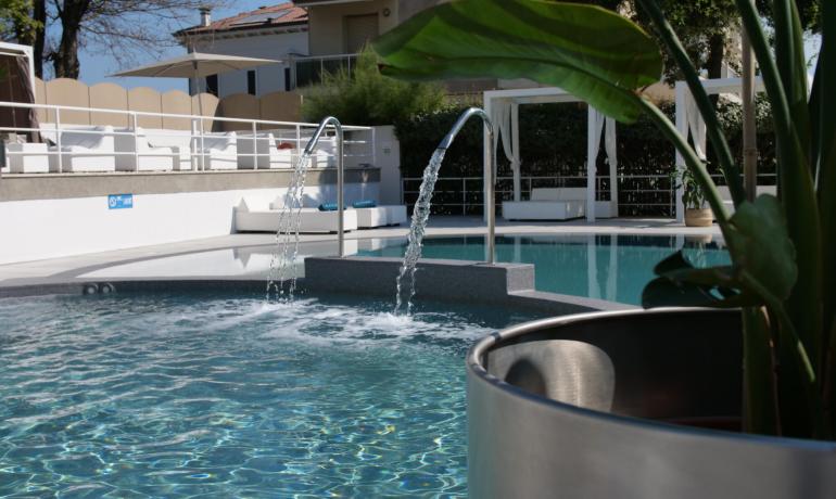 Piscina con getti d'acqua, lettini e vegetazione.