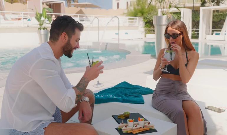 Due persone rilassate a bordo piscina bevono cocktail e mangiano snack.