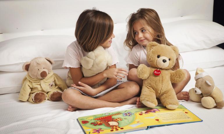 Due bambine sorridenti con orsacchiotti leggono insieme un libro colorato sul letto.