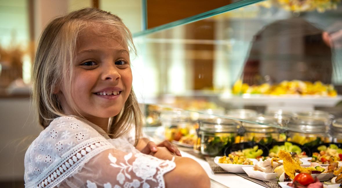 Bambina sorridente vicino a un buffet di cibo, indossa un vestito bianco.