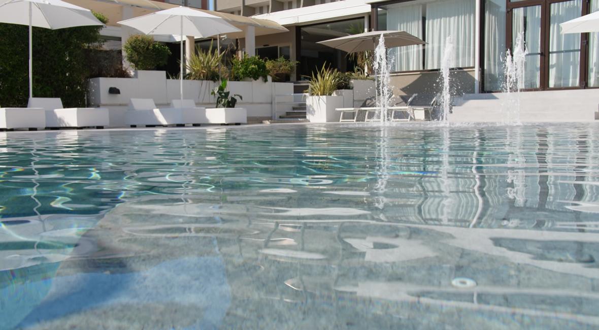 Piscina con lettini bianchi e ombrelloni, fontane d'acqua e vegetazione.