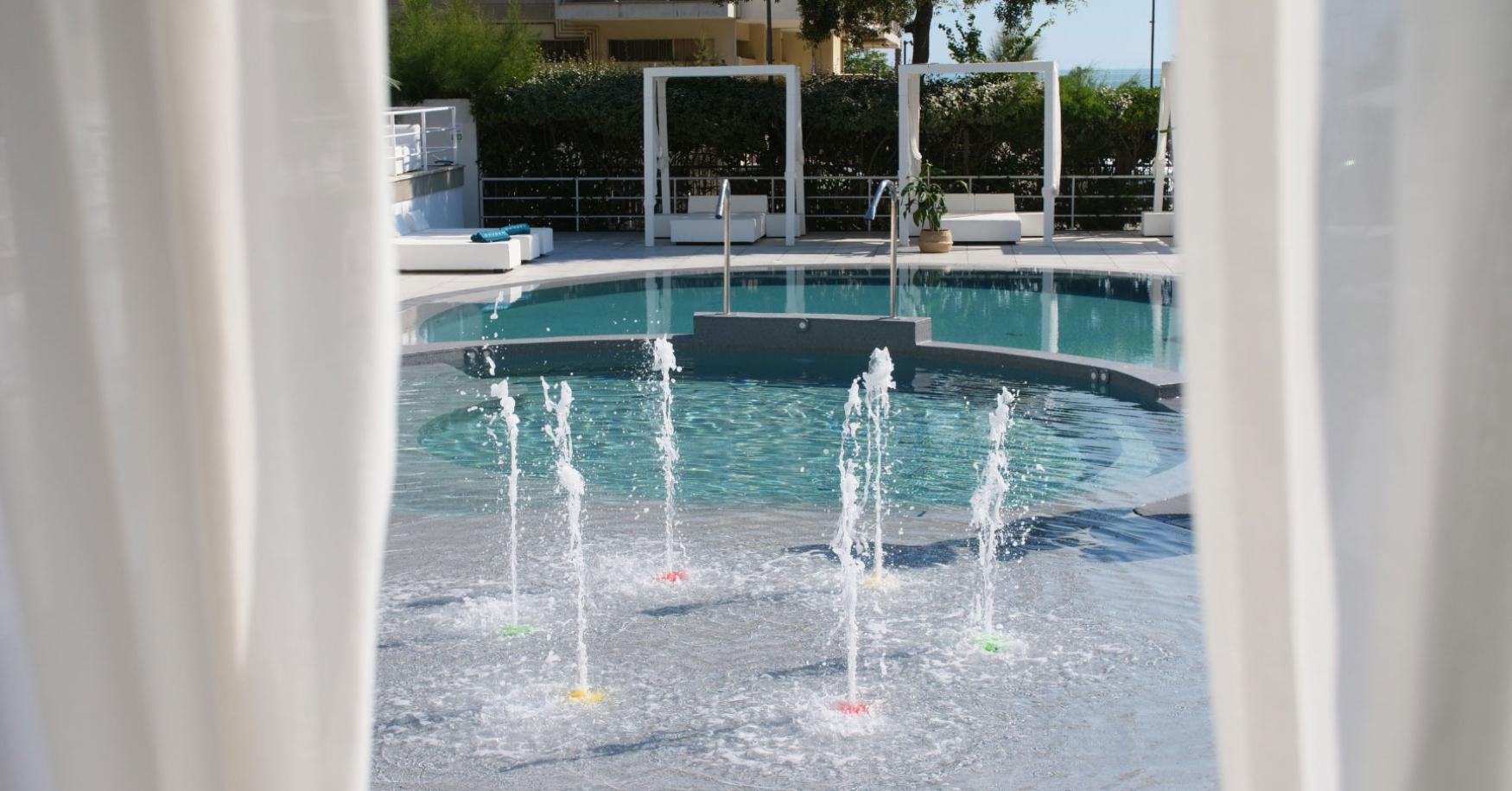 Piscina con fontane d'acqua e area relax, vista attraverso tende bianche.