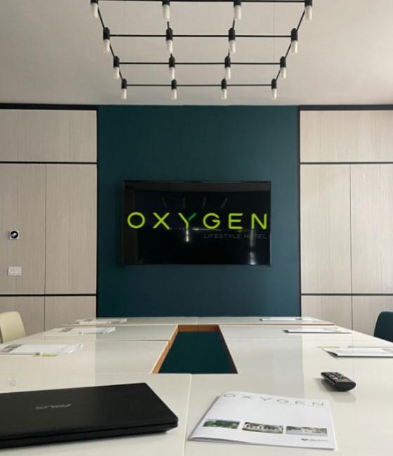 Sala riunioni moderna con schermo, tavolo bianco e logo Oxygen Lifestyle Hotel.